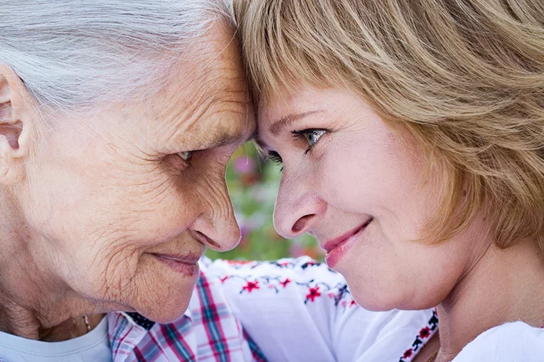 Elderly mother and adult daughter looking into each other's eyes, smiling with a sense of support.